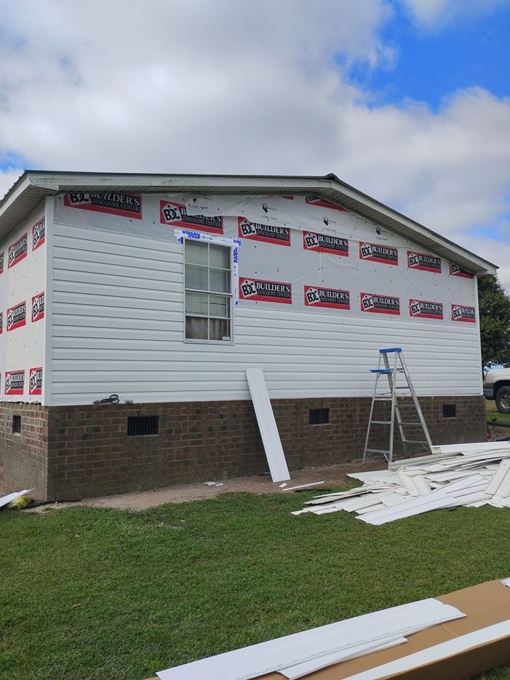 A house under construction with partial white siding, exposed sheathing, a single window, and a ladder on the grass.