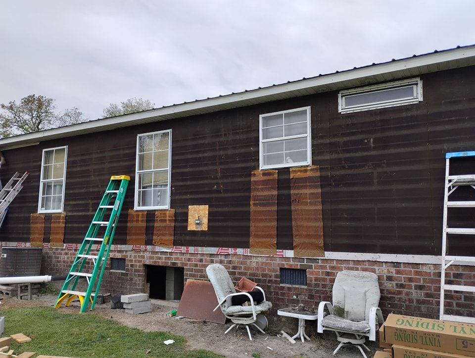 A house under renovation with dark siding, a brick foundation, a green ladder, two windows, and boxes of new vinyl siding.