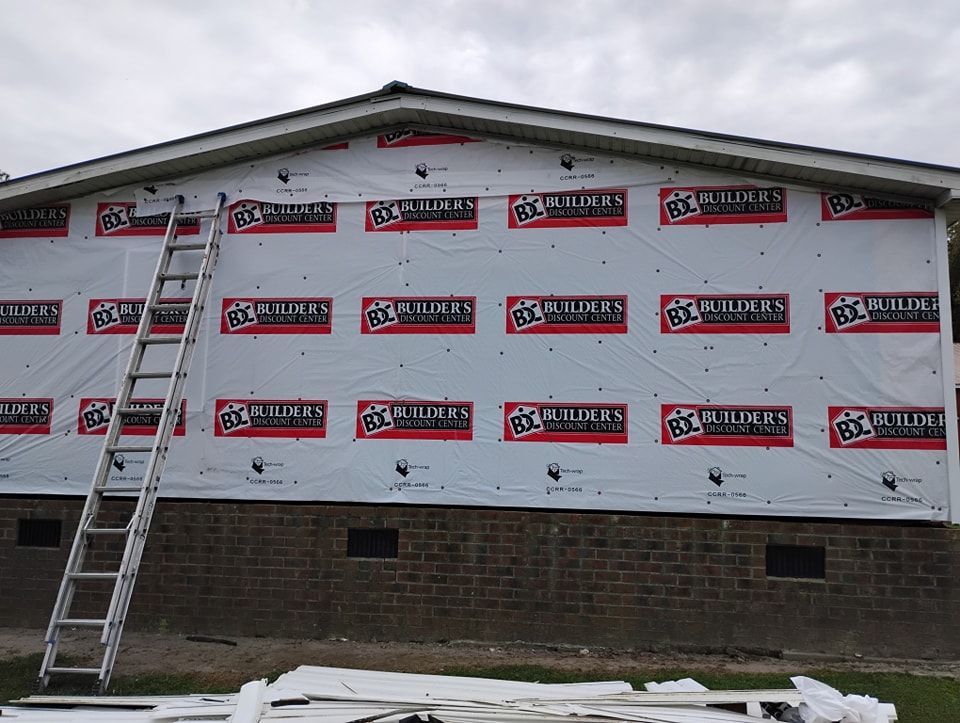 A house exterior undergoing renovation, featuring white house wrap over the walls, a brick foundation, and a metal ladder.