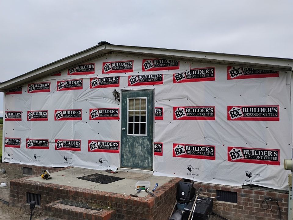A house exterior under construction, covered in white vapor barrier sheeting with red and black brand labels.