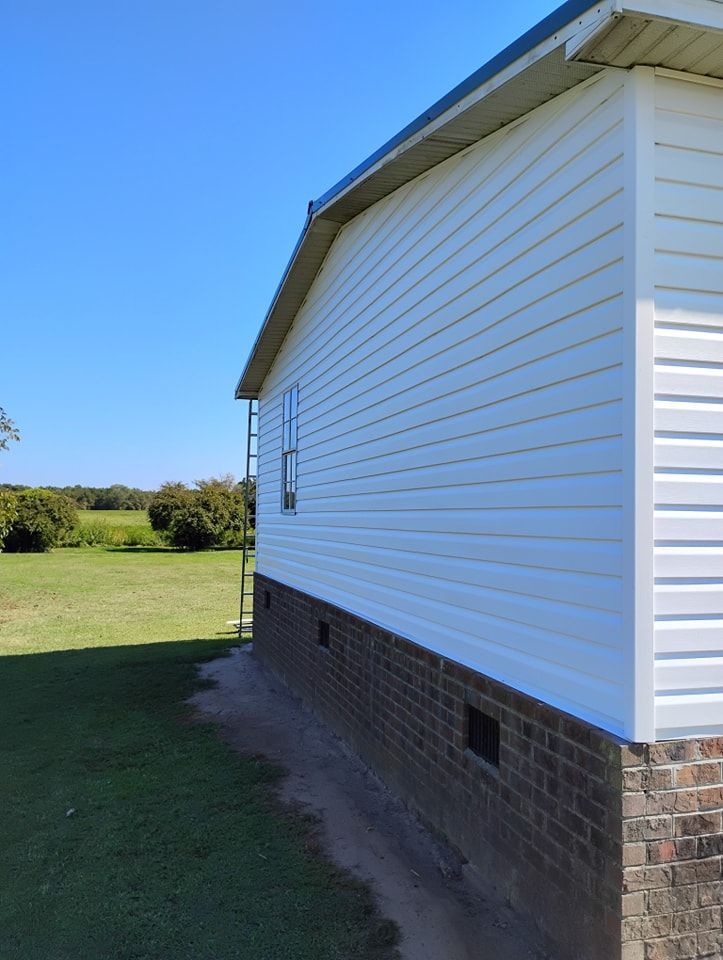 Side view of a house with white vinyl siding, a brick foundation, and a ladder against the wall in a grassy field.