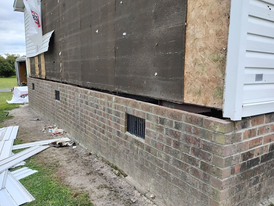 A side view of a house foundation with exposed brick, wall sheathing, and discarded siding on the ground nearby.