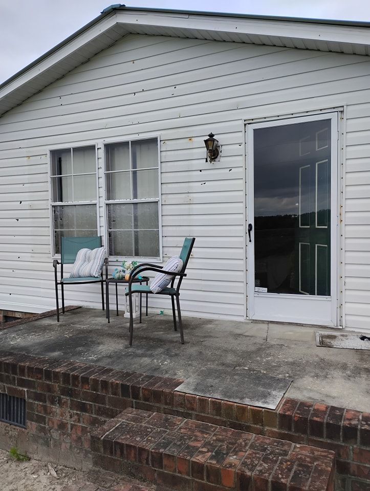 A white, horizontal-sided home features a brick stoop, two chairs and a small table on a concrete porch, and a glass door.