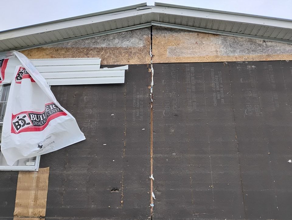 A house exterior wall showing exposed dark sheathing, a central vertical gap, and loose building wrap under the roofline.