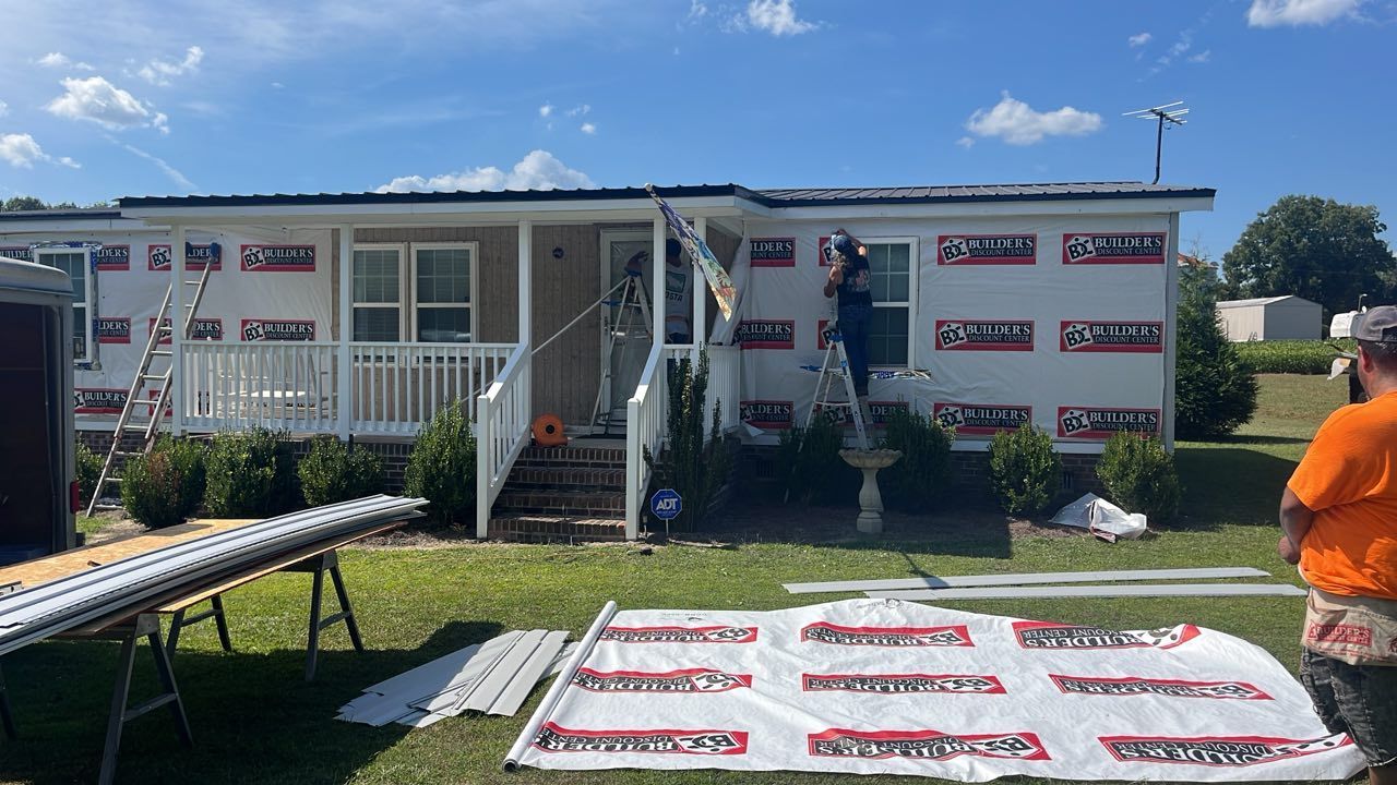 Workers install house wrap and siding on the exterior of a white manufactured home on a sunny day.