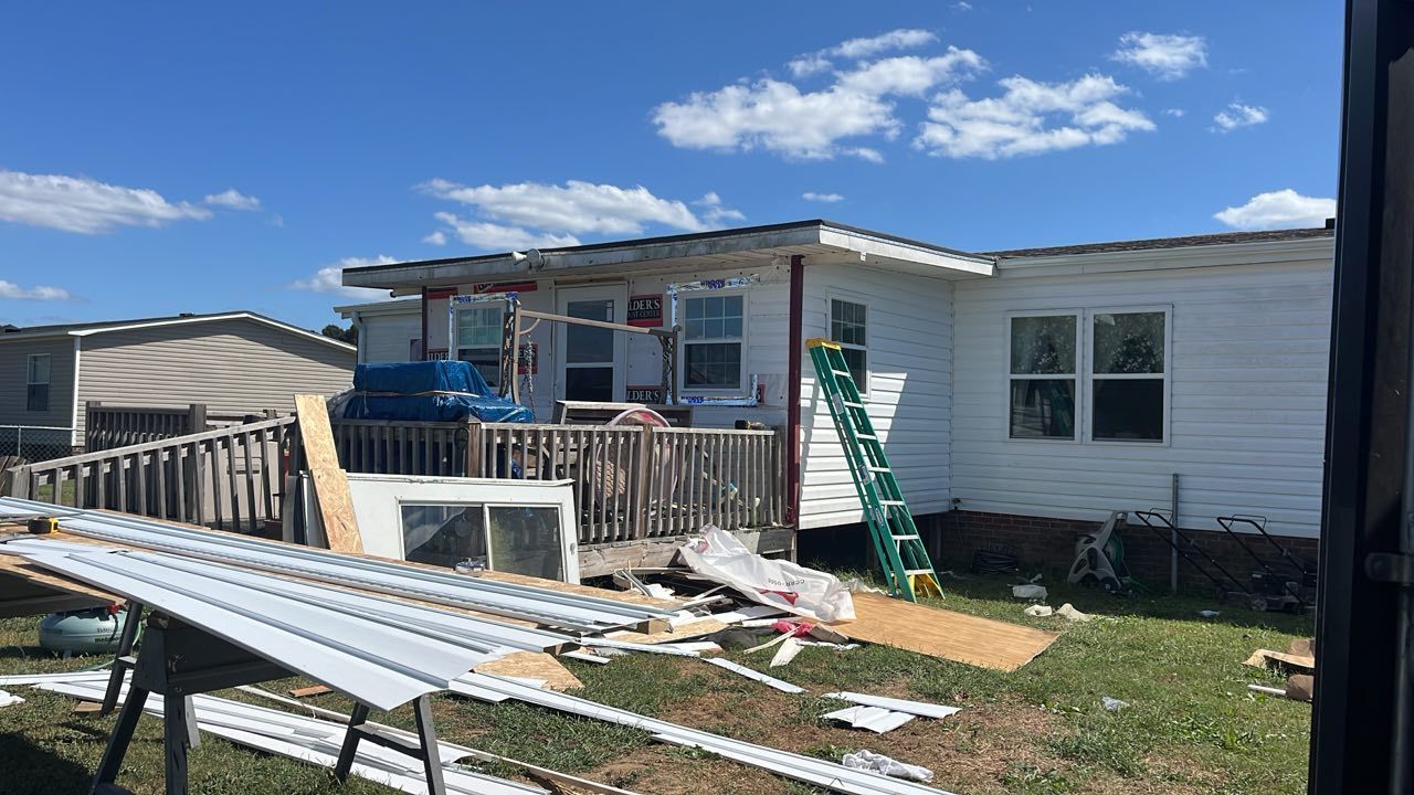 A white house under repair with a green ladder, wooden deck, and stacks of siding in the front yard.