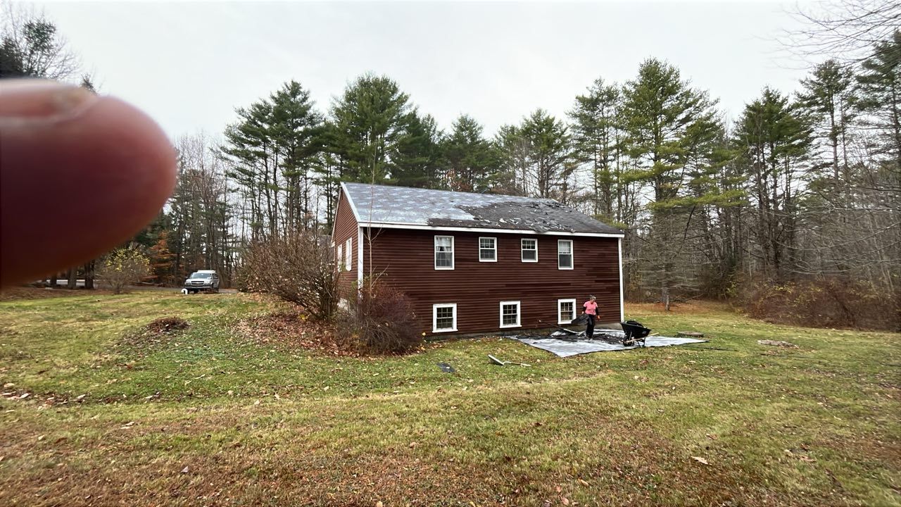 A brown, two-story house stands in a grassy field bordered by trees, with people working on a project near the foundation.