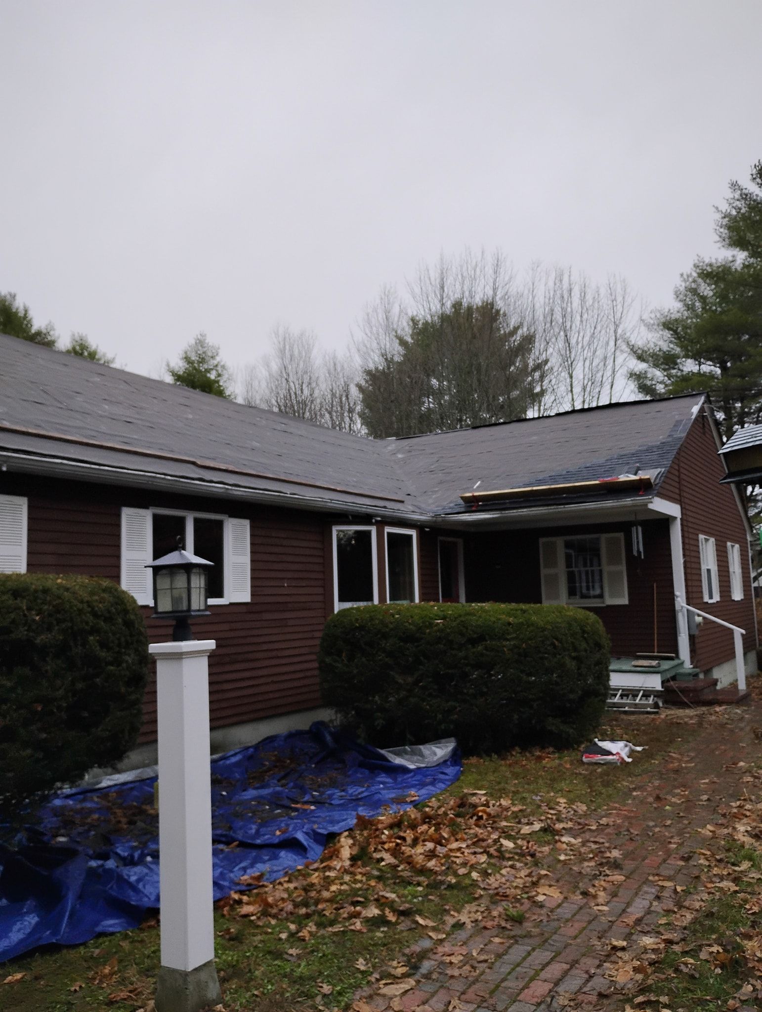 A brown house with a damaged roof section, blue tarp on the ground below, and white window shutters on an overcast day.