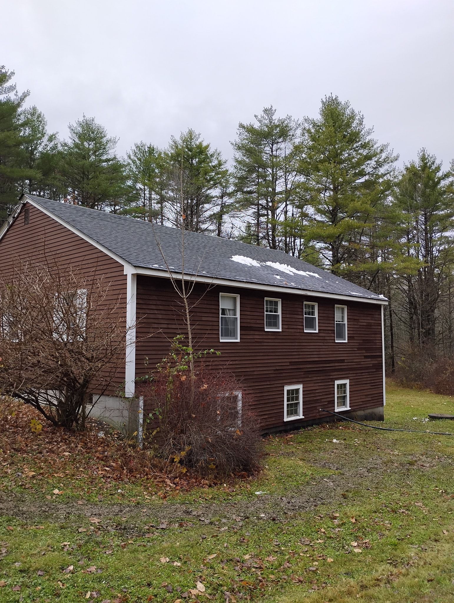 A brown, two-story house with a shingled roof stands amidst a wooded landscape under an overcast sky.