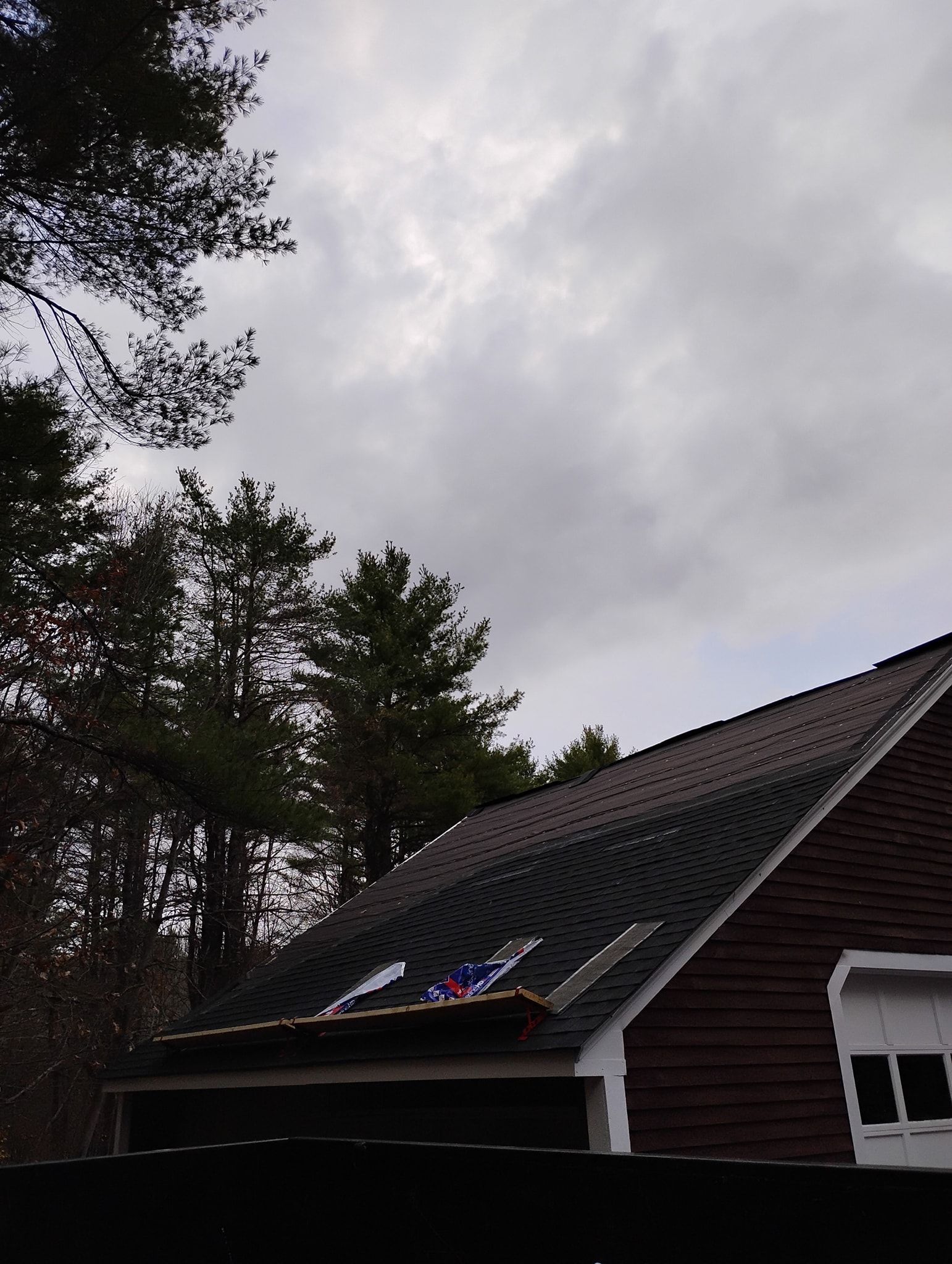 A house roof with shingles partially missing, showing exposed wood and temporary blue tarp patching under a cloudy sky.