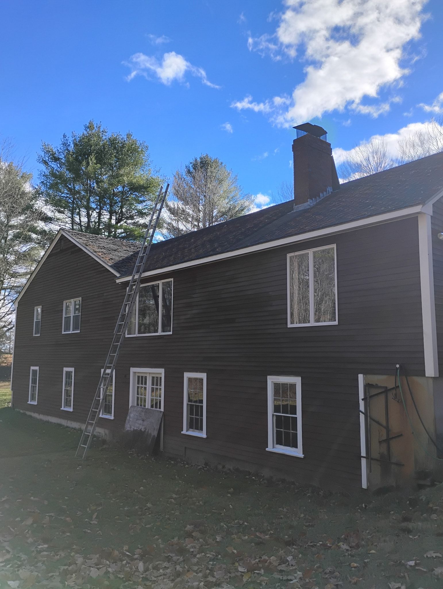 A two-story dark-sided house with white trim and a chimney, viewed from a grassy yard under a blue sky with clouds.
