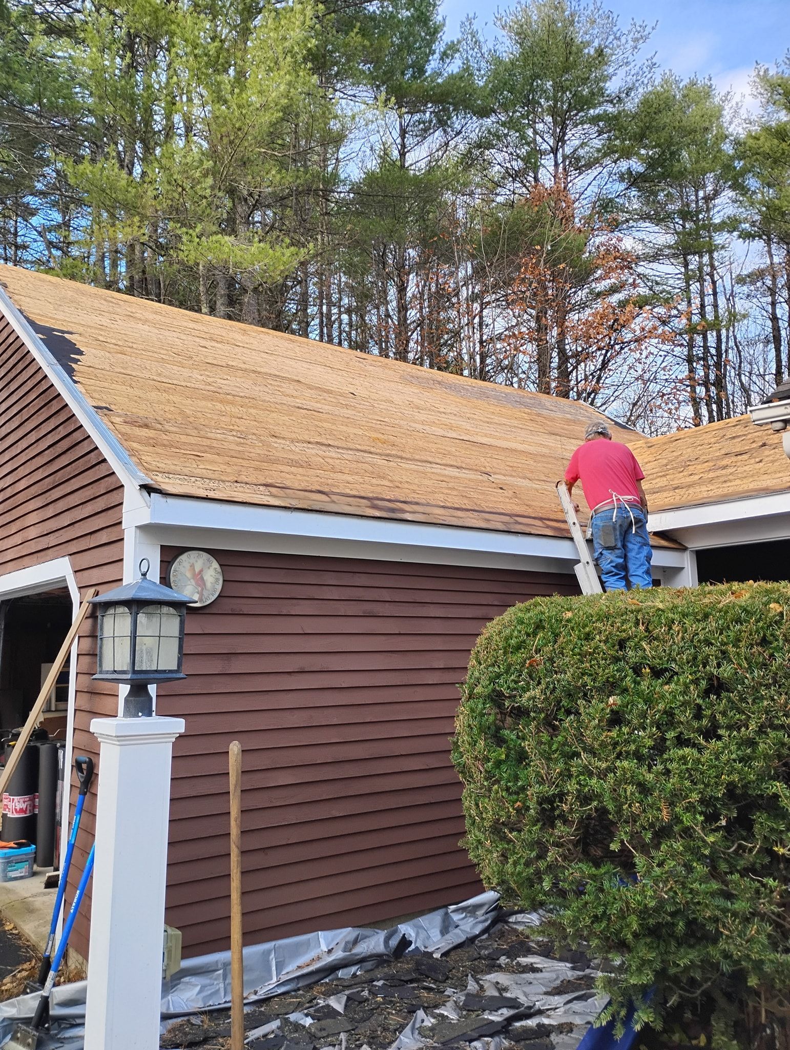 A person in a red shirt works on the roof of a brown house with dark brown siding, viewed from outside near a bush.
