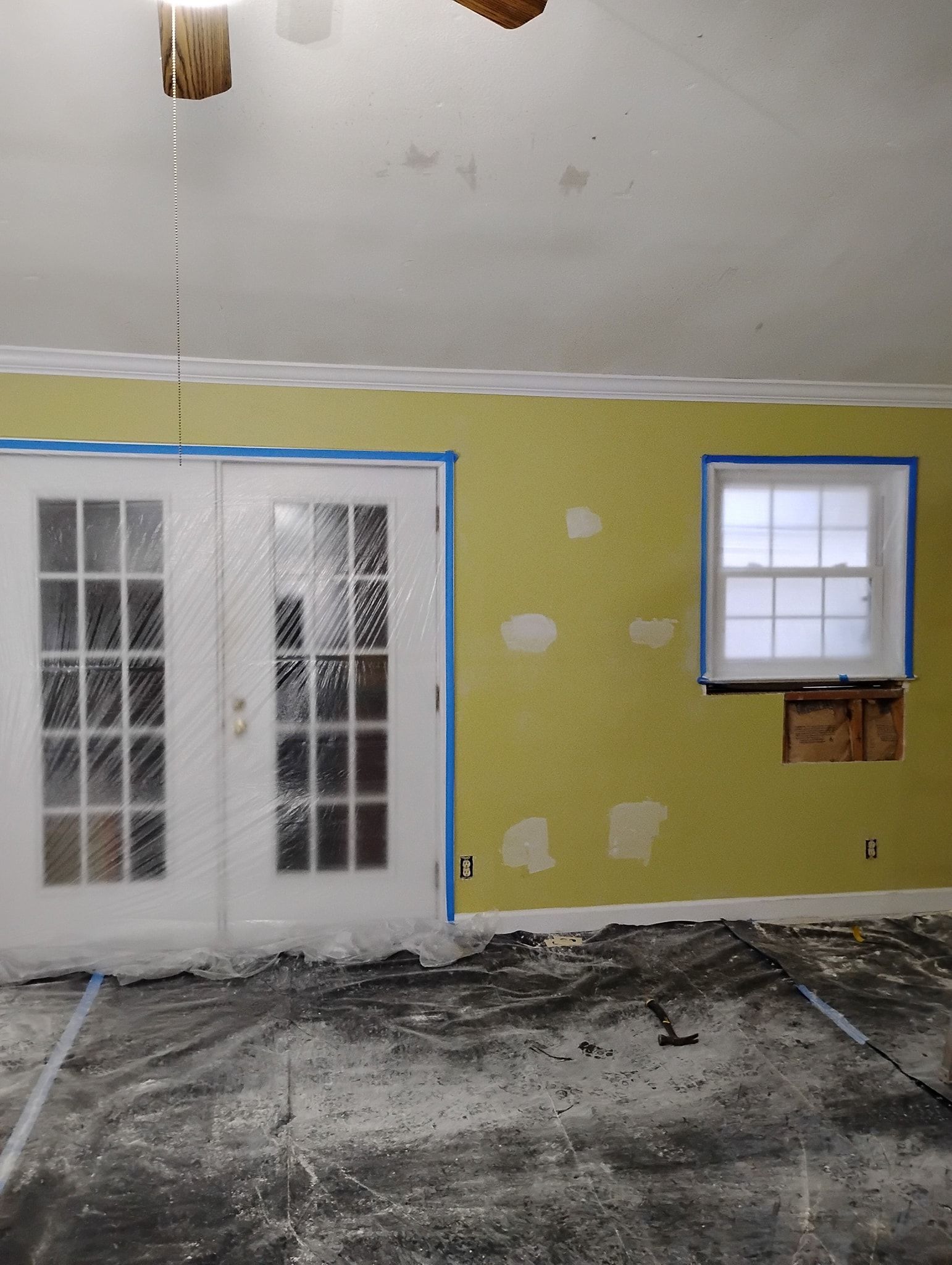 A room under renovation with a yellow wall featuring patch repairs, double glass doors, a window, and a dusty floor.