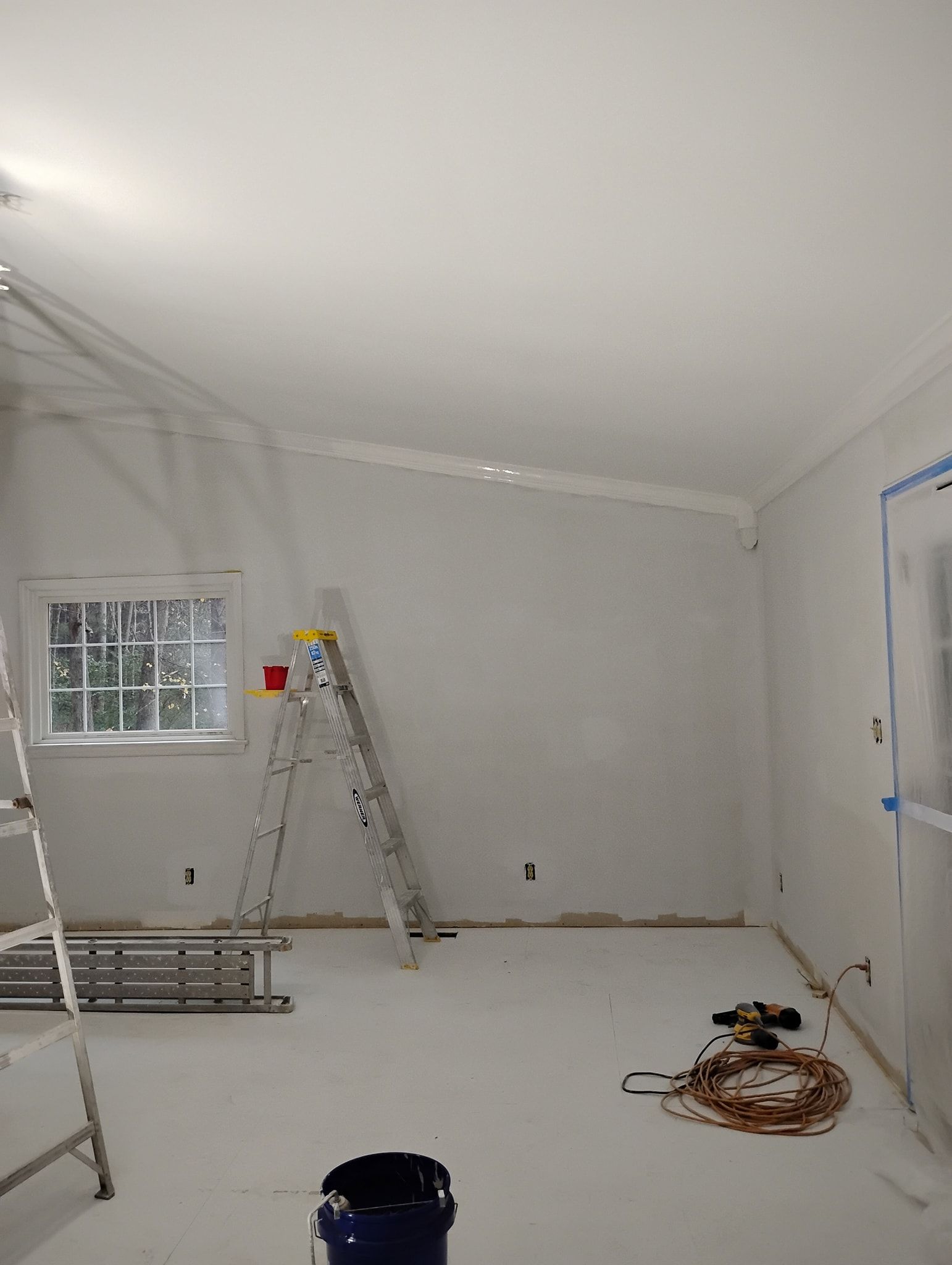 A room under renovation with white walls, a step ladder, a paint bucket, and electrical supplies on a drop cloth.