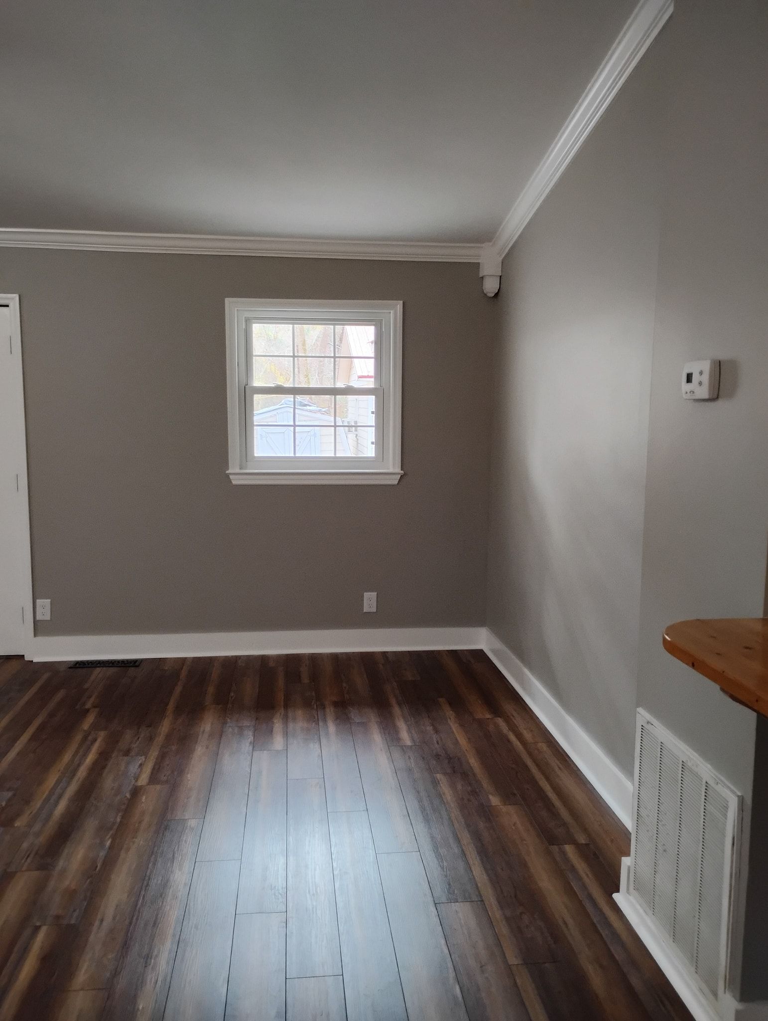 A room with grey walls, dark wood-look plank flooring, white crown molding, baseboards, and a single window.