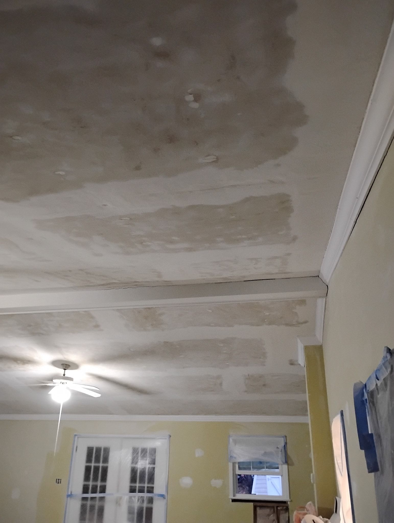 Ceiling with a large, dark water stain and visible joint lines against a beige wall in a room with a ceiling fan.