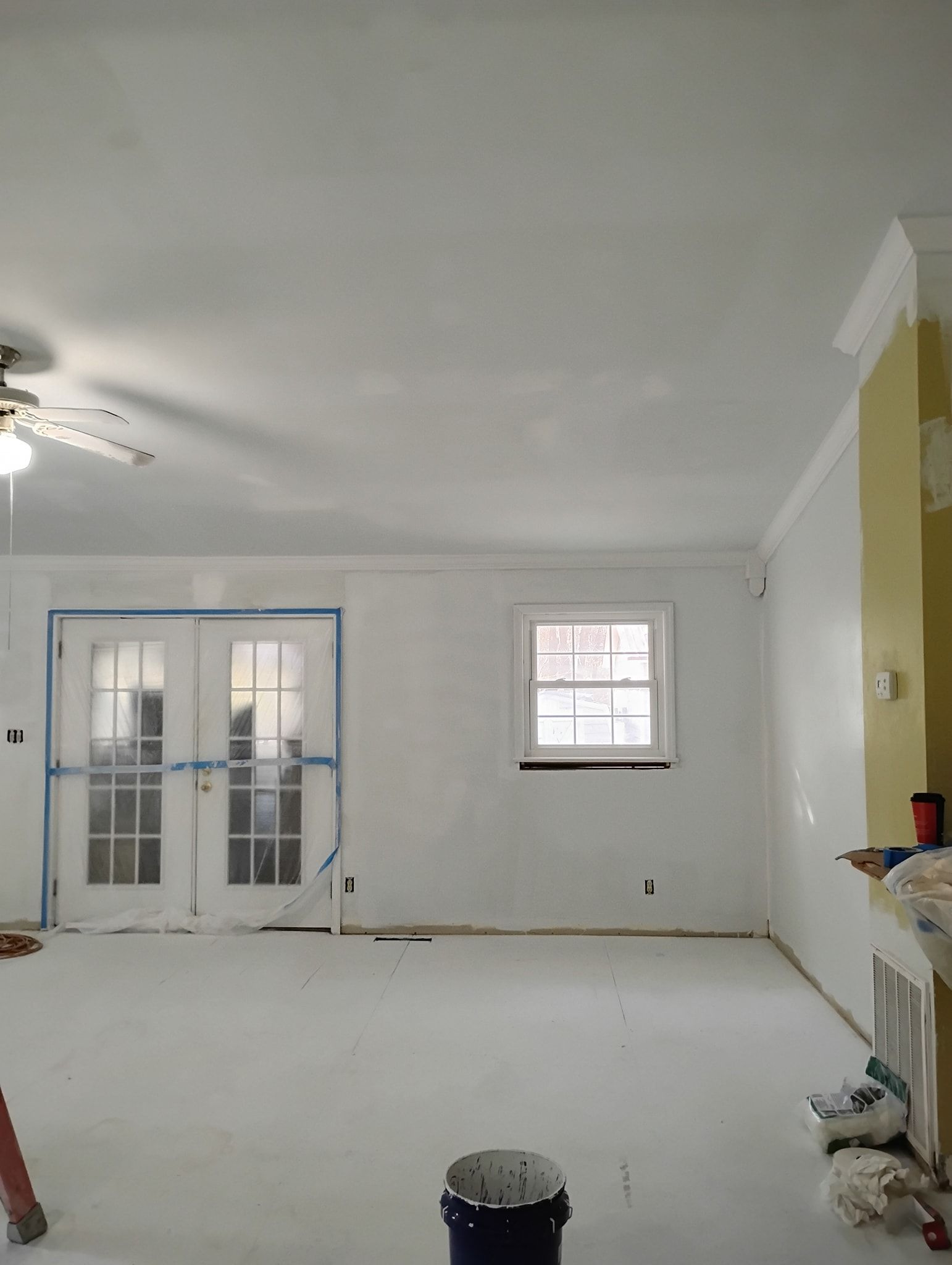 A room undergoing renovation with freshly painted white walls and ceiling, a window, French doors, and a yellow wall section.