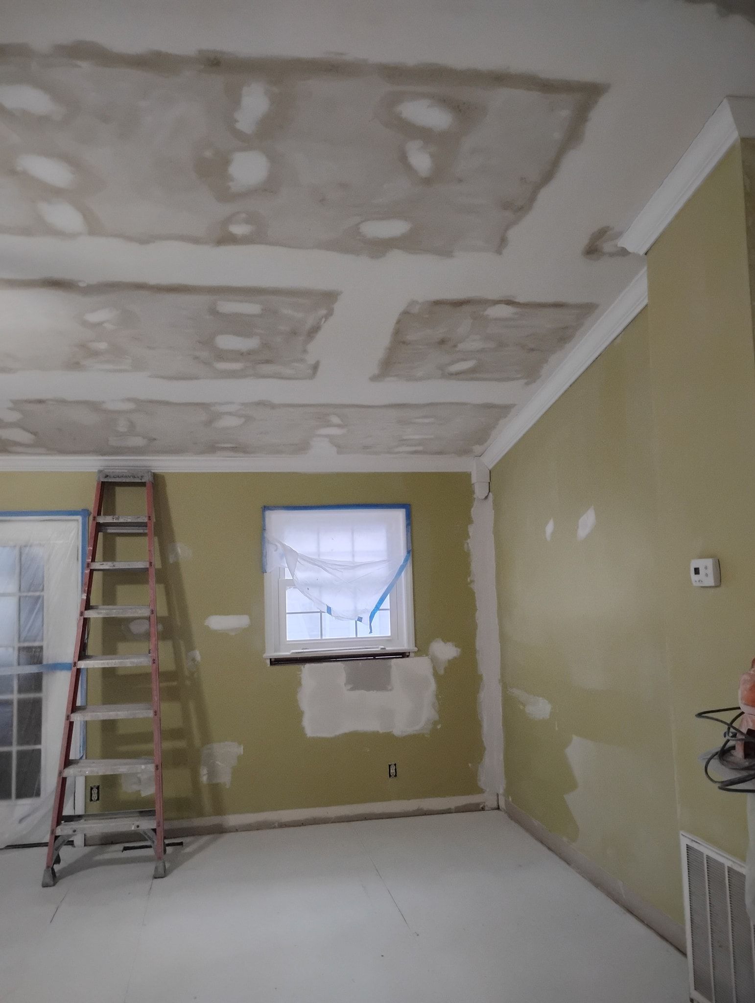 An interior room under renovation with a patch-worked ceiling, taped windows, and a tall ladder against a yellow wall.
