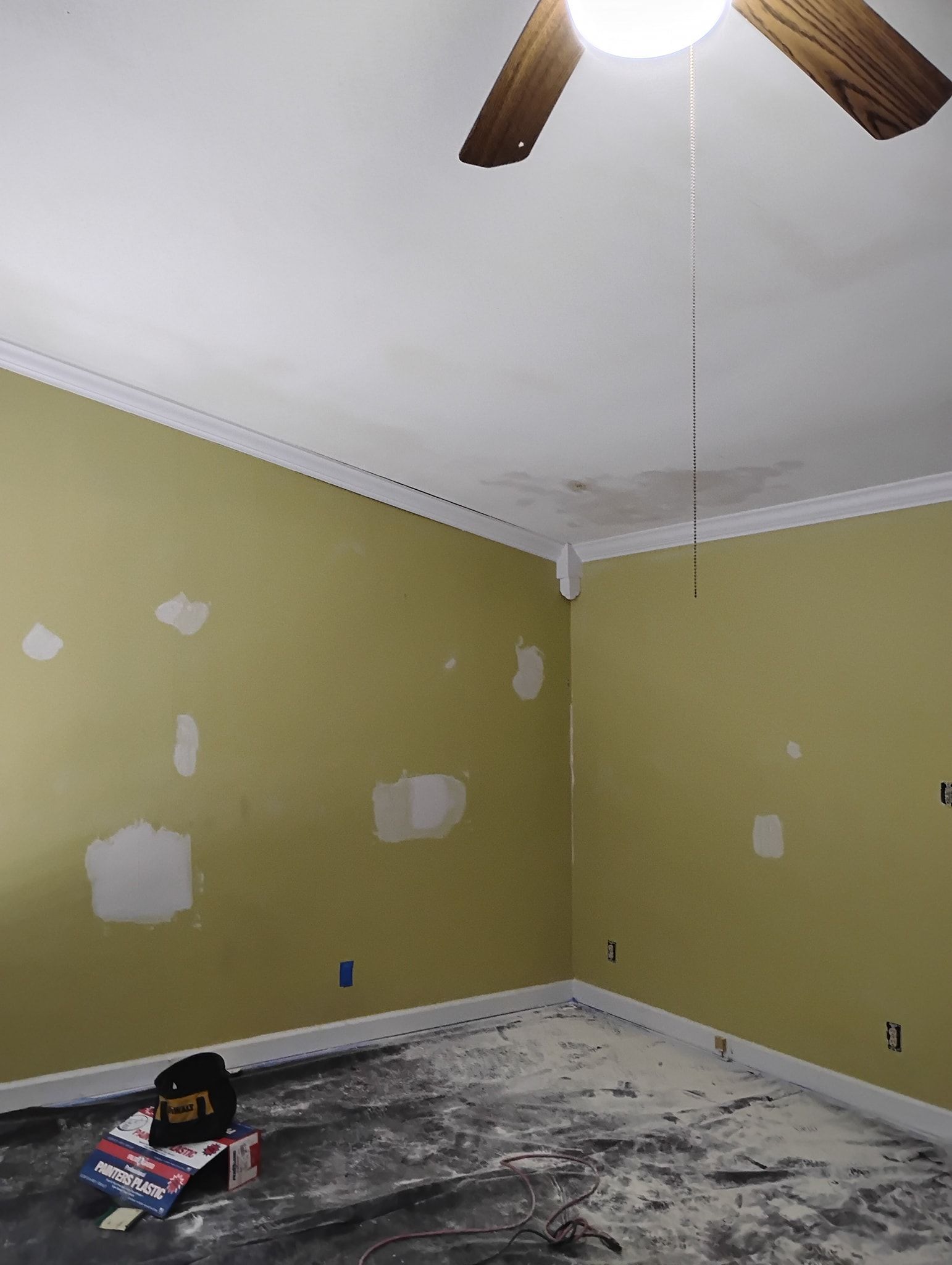 A room under renovation with yellow walls showing filled drywall patches and a ceiling with a fan and water stains.