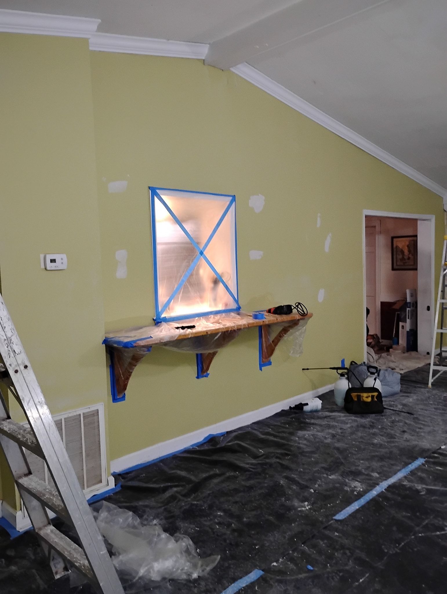 A room with light green walls being prepped for painting, featuring a taped-off window, wooden brackets, and a ladder.