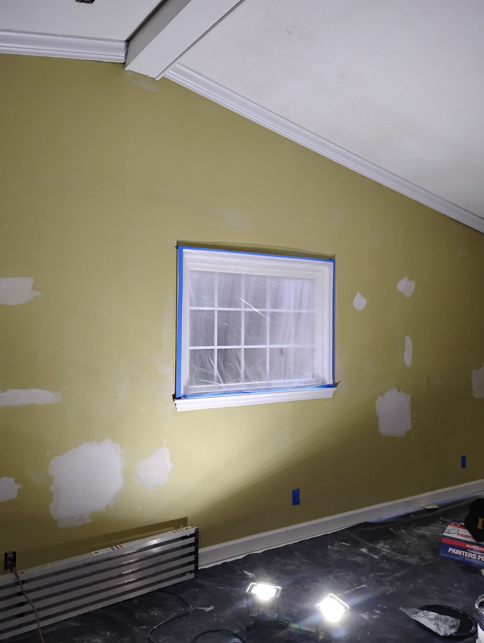 An interior wall with patches of white filler, a window covered in blue painter's tape, and a baseboard heater.