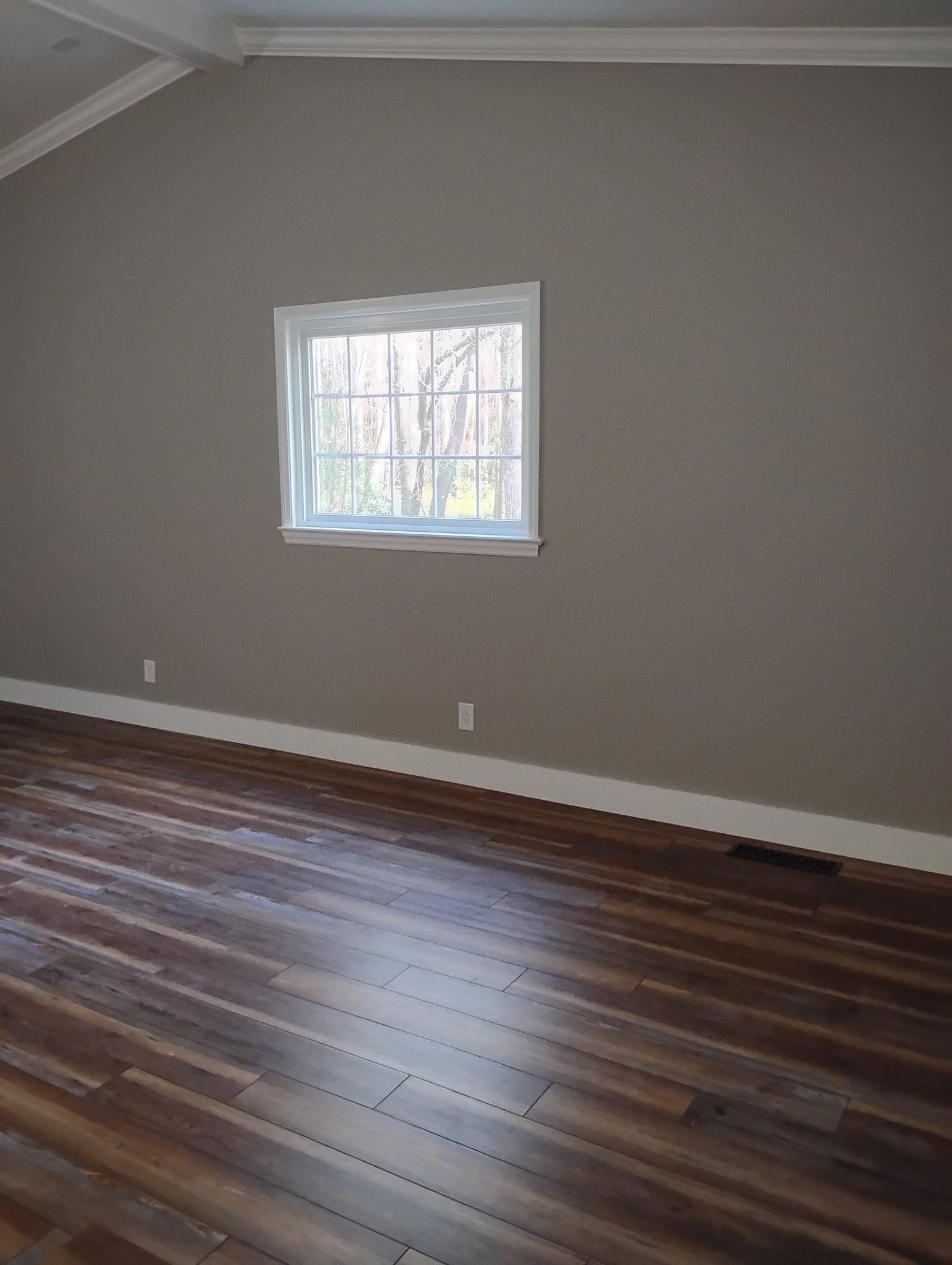 A room with brown walls, white trim, a single square window centered on the wall, and dark wood plank flooring.