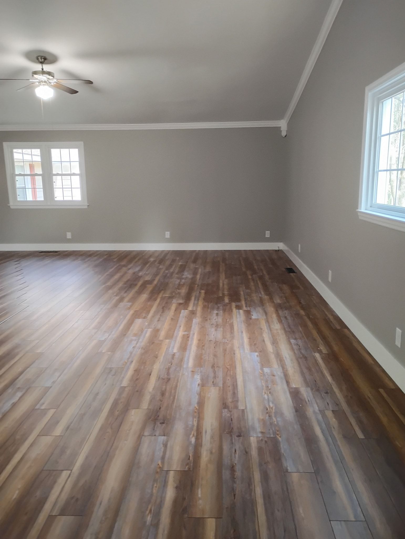 A large, empty room with grey walls, white trim, wood-look flooring, and a ceiling fan.