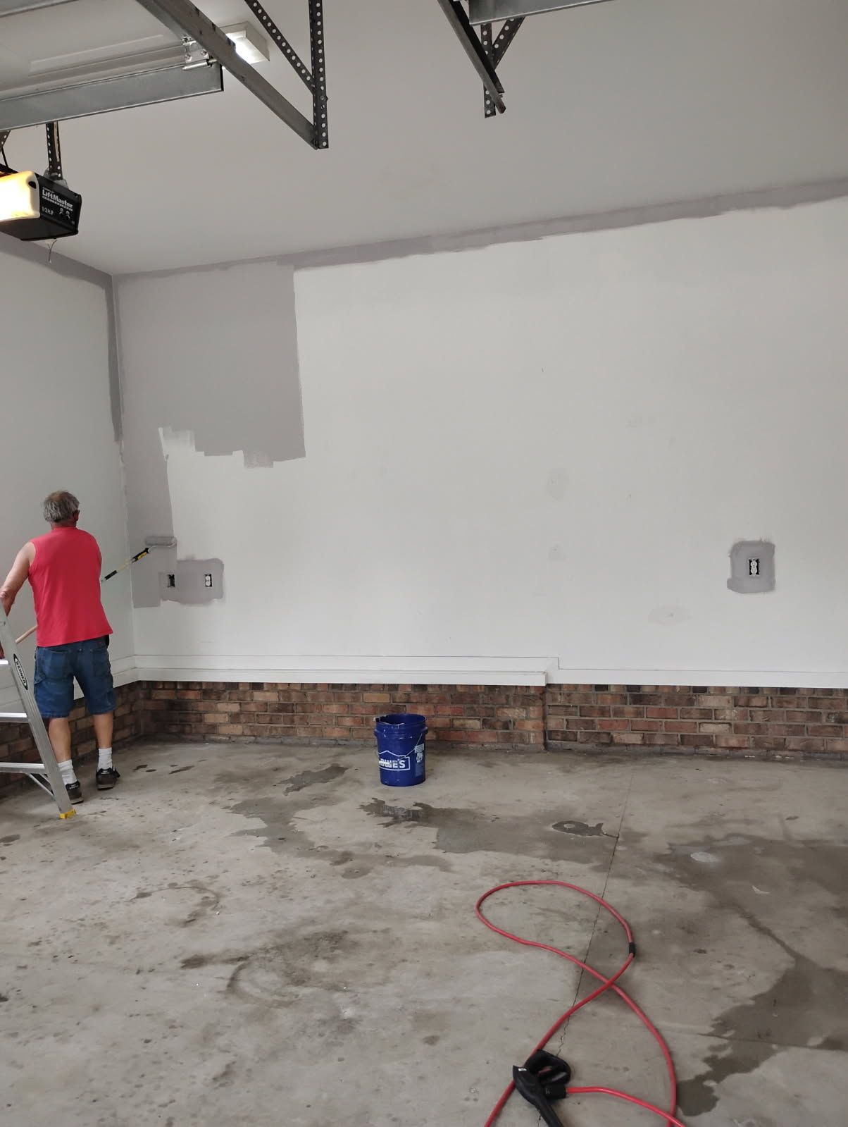 Person painting a garage wall white, with exposed brick below and a paint bucket on the floor.