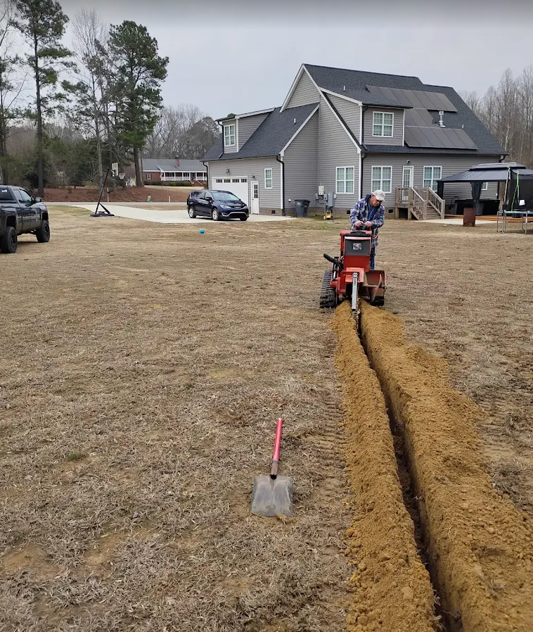 Worker operating a trencher, cutting a narrow trench across a yard near a house.