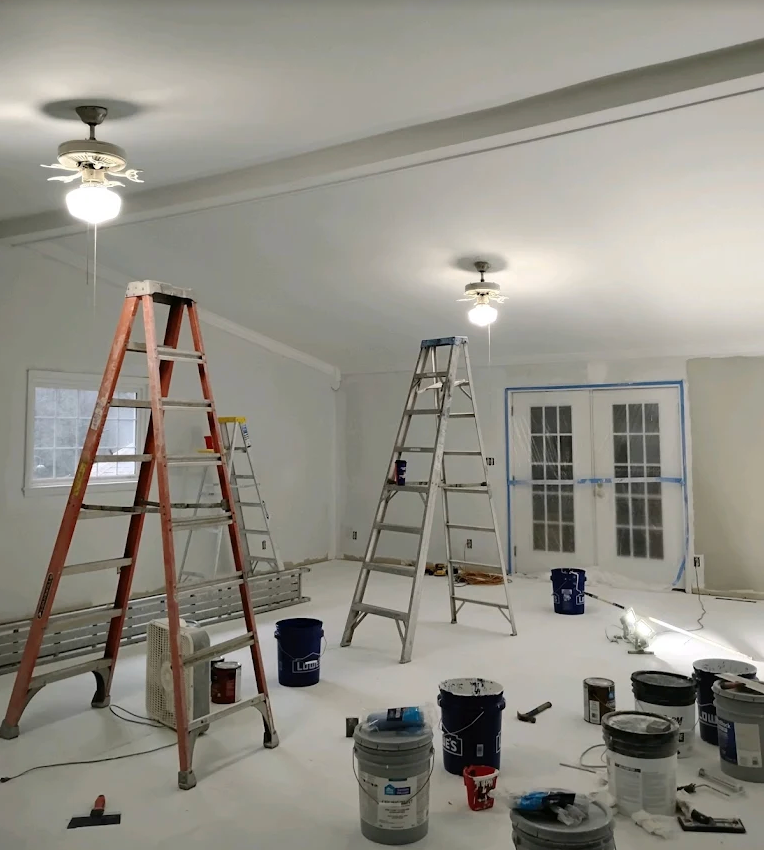 Interior room under renovation with ladders, paint cans, and ceiling fans on a covered floor.