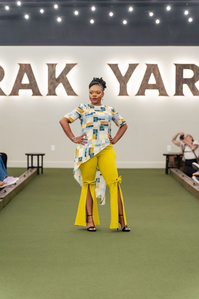 Woman in yellow pants and patterned top on a runway, hands on hips.