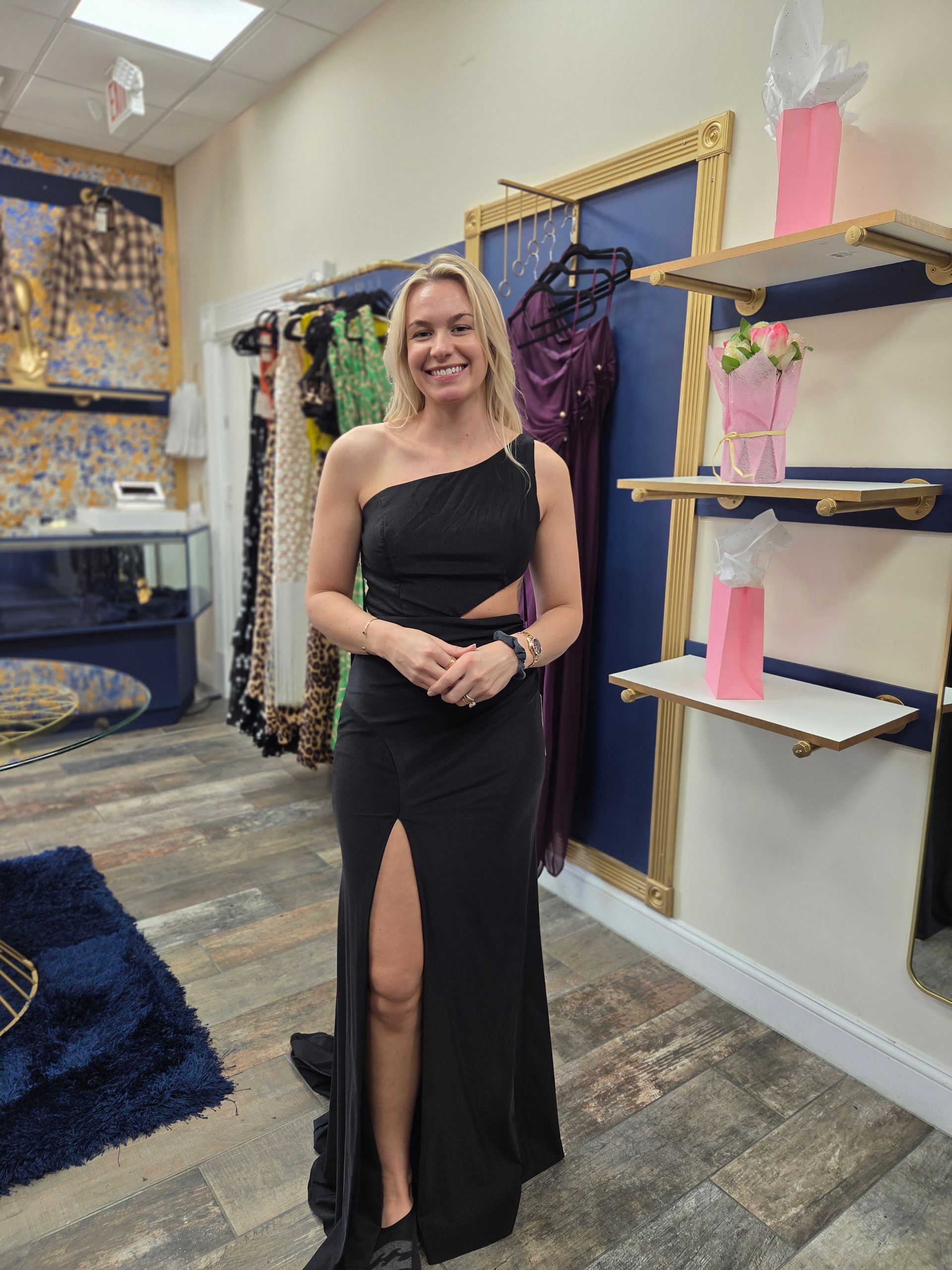 Woman in black formal dress, posing in a clothing store. She's smiling, dress has a thigh slit and cutout.