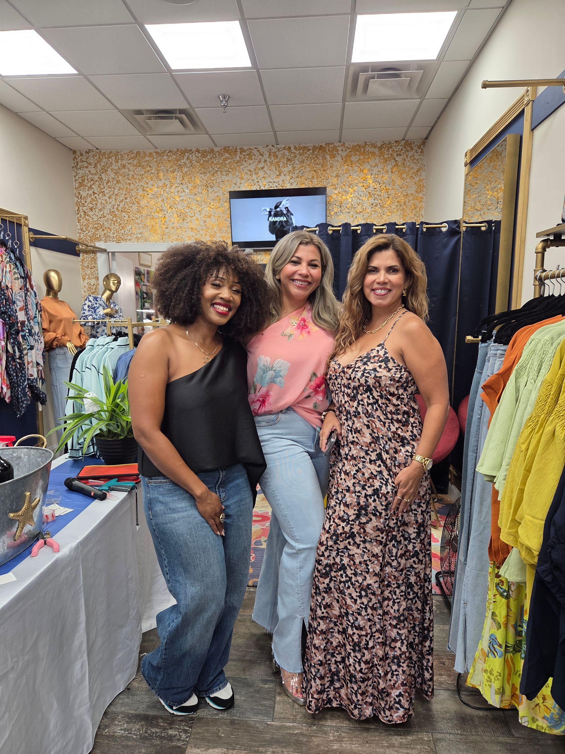 Three women smile, posing in a clothing store. Two are in dresses, one in top and jeans. Store displays clothing.