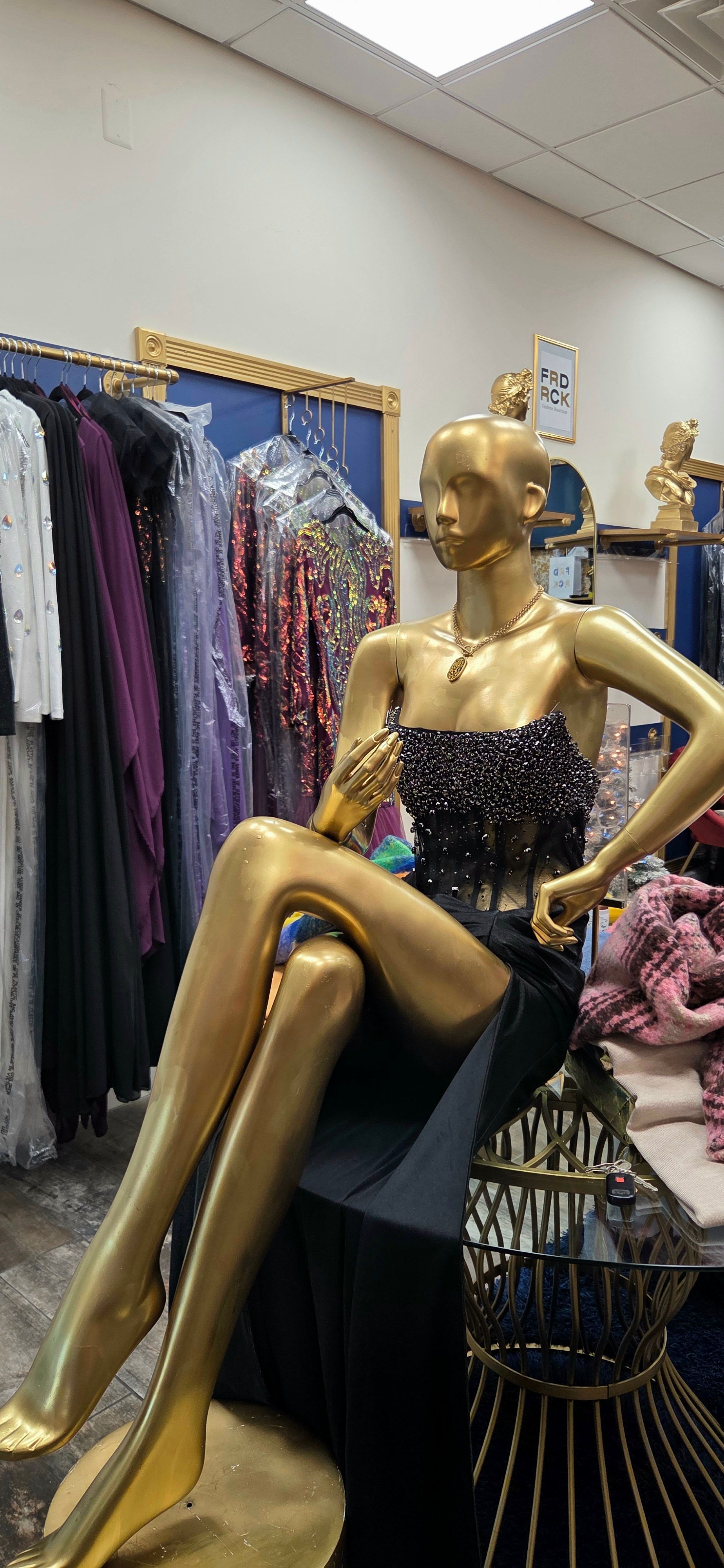 Golden mannequin in a black dress with a jeweled bodice, sitting indoors near racks of clothing.