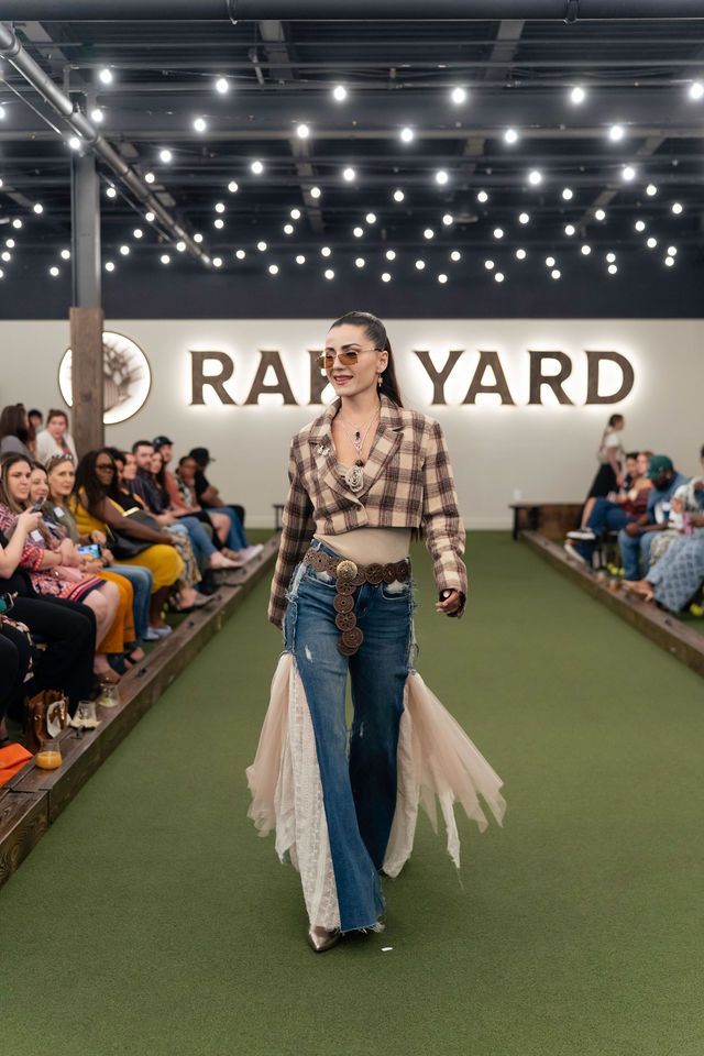 Woman walks runway at a Raft Yard fashion show. She wears a plaid crop top, jeans, and sunglasses.