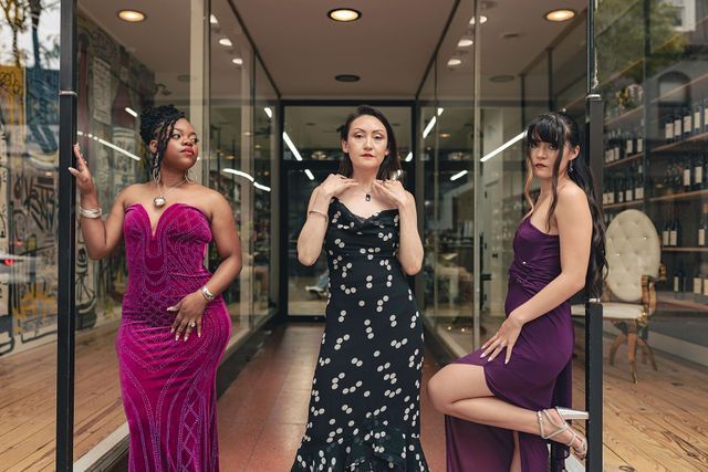 Three people in formal dresses pose in front of a glass storefront.