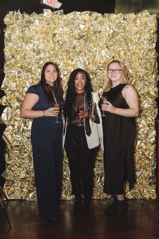 Three people pose in front of a gold backdrop. Each holds a drink. One wears a black outfit, the others wear dark-colored outfits.