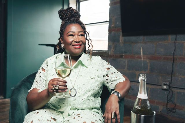 Woman holding wine, wearing green lace, smiling in a room with brick wall and champagne.