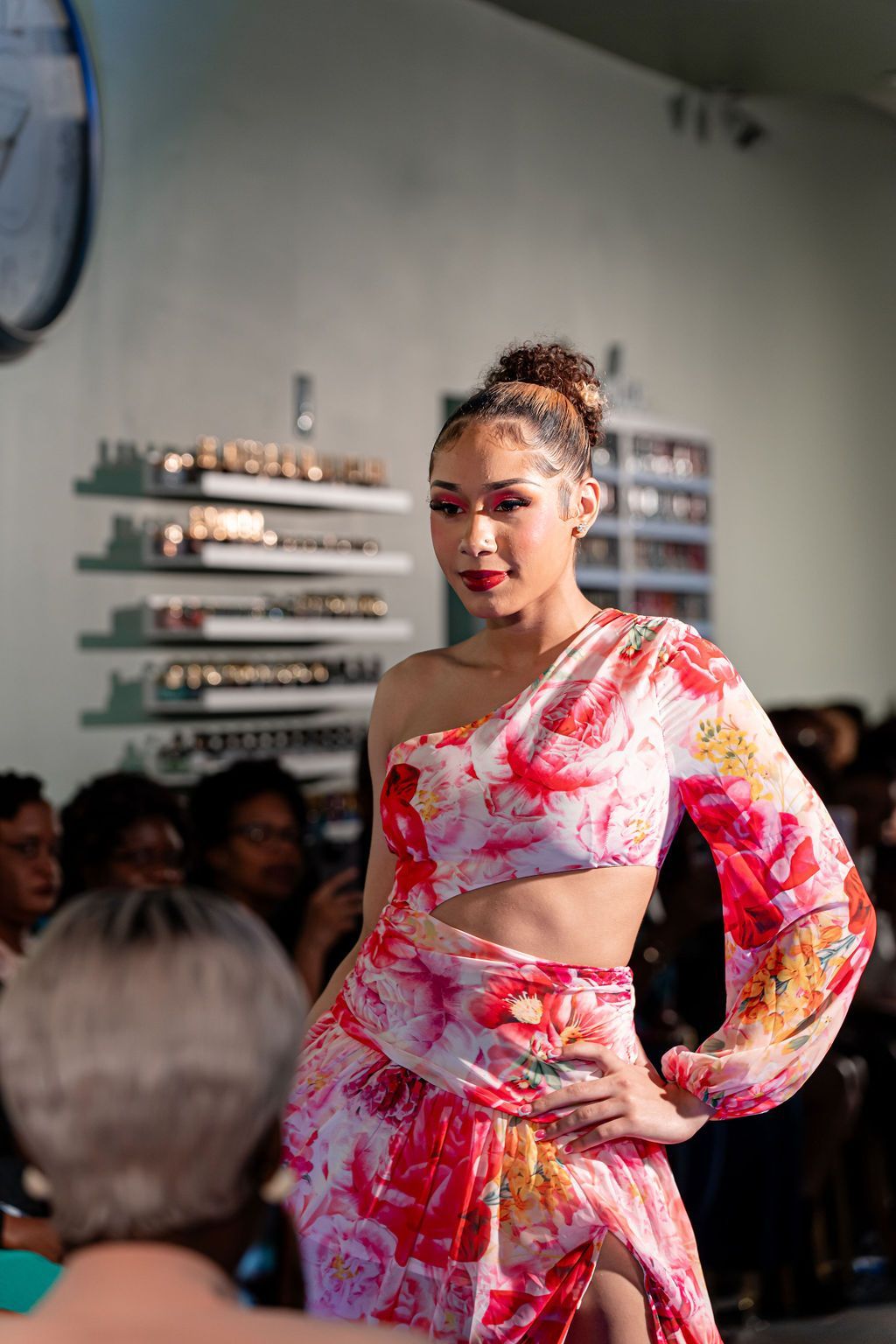Woman in floral one-shoulder dress on a runway, hands on hip, with audience behind her.