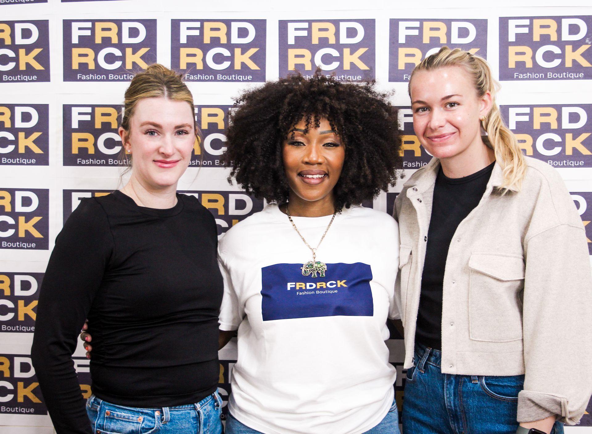 Three women pose in front of a backdrop with logos. Woman in center wears a white tee with the word