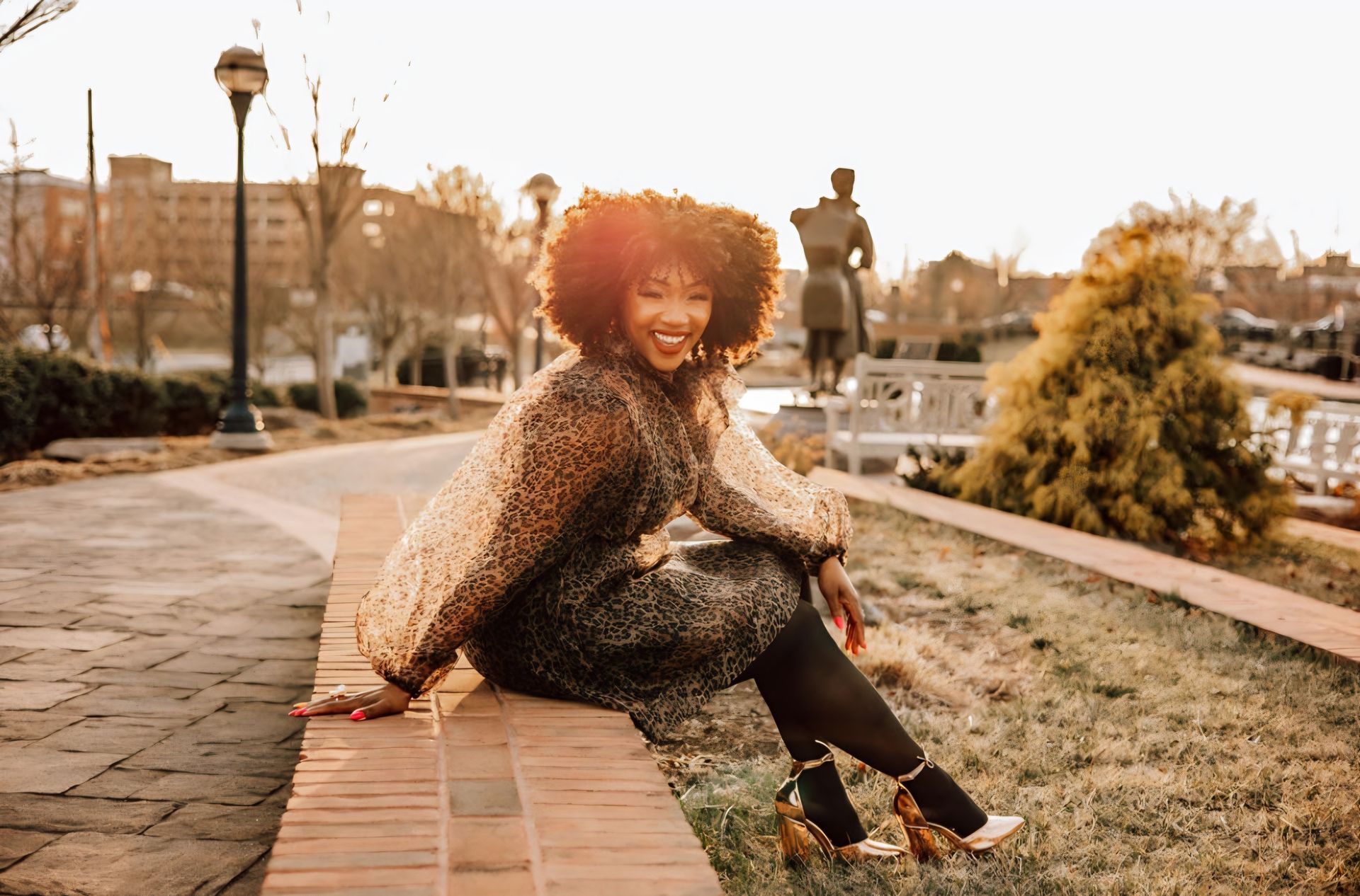Woman with large afro smiling, seated on brick wall in park, wearing sparkly dress, gold heels; sunlight.