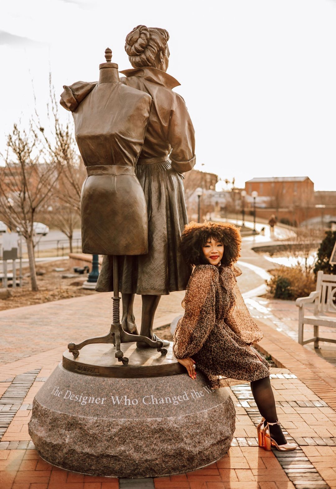 Woman poses next to a statue of a seamstress and a dress form in a town square; she wears a leopard print dress.