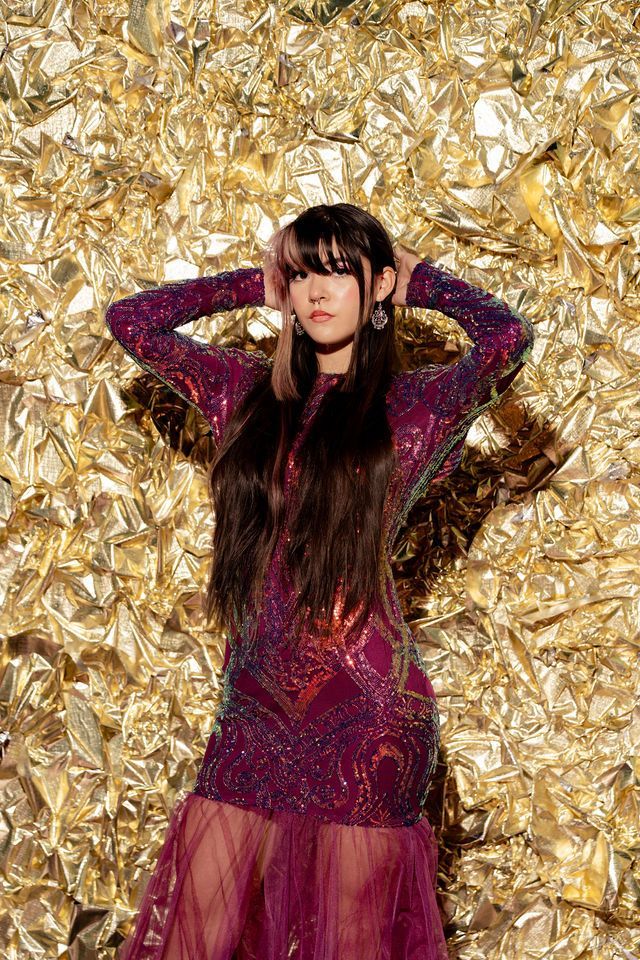 Woman in sequined purple dress poses with hands behind head, against gold textured background.