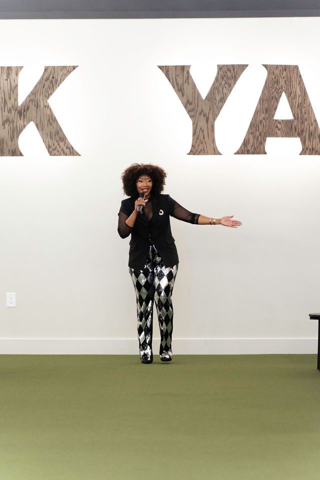Woman speaking, gesturing, standing on green carpet, in front of a white wall with