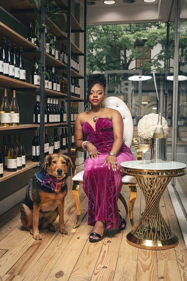 Woman in pink dress sits with dog in wine shop. Gold table with wine glass. Wooden floors, shelves of wine.
