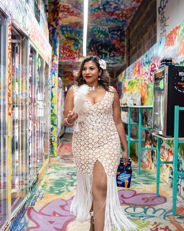 Woman in white dress with feathered boa, holds beer, walks through colorful graffiti hallway.