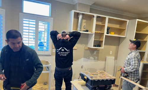 Three men installing kitchen cabinets in a remodeled room. One measures, one adjusts, one looks up.