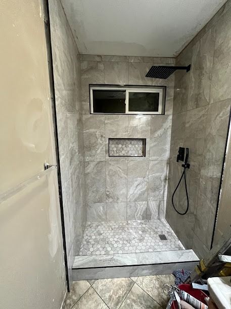 Newly tiled shower stall with a dark shower head and window, gray tiles, and a recessed shelf.