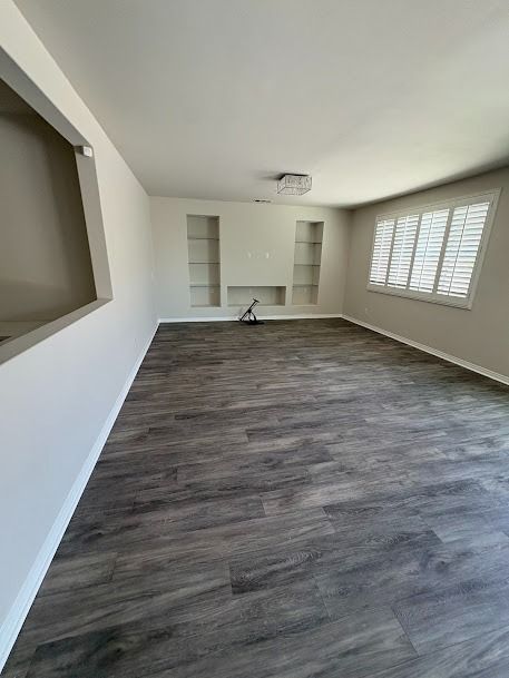 Empty living room with grey wood-look flooring, white walls, built-in shelves, and window with shutters.