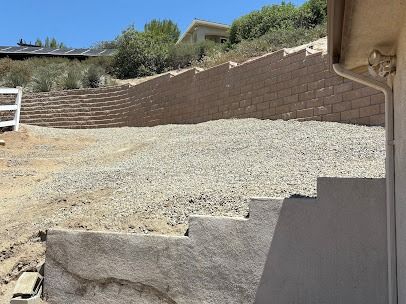 A gravel yard with a retaining wall, steps, and a house, on a sunny day.